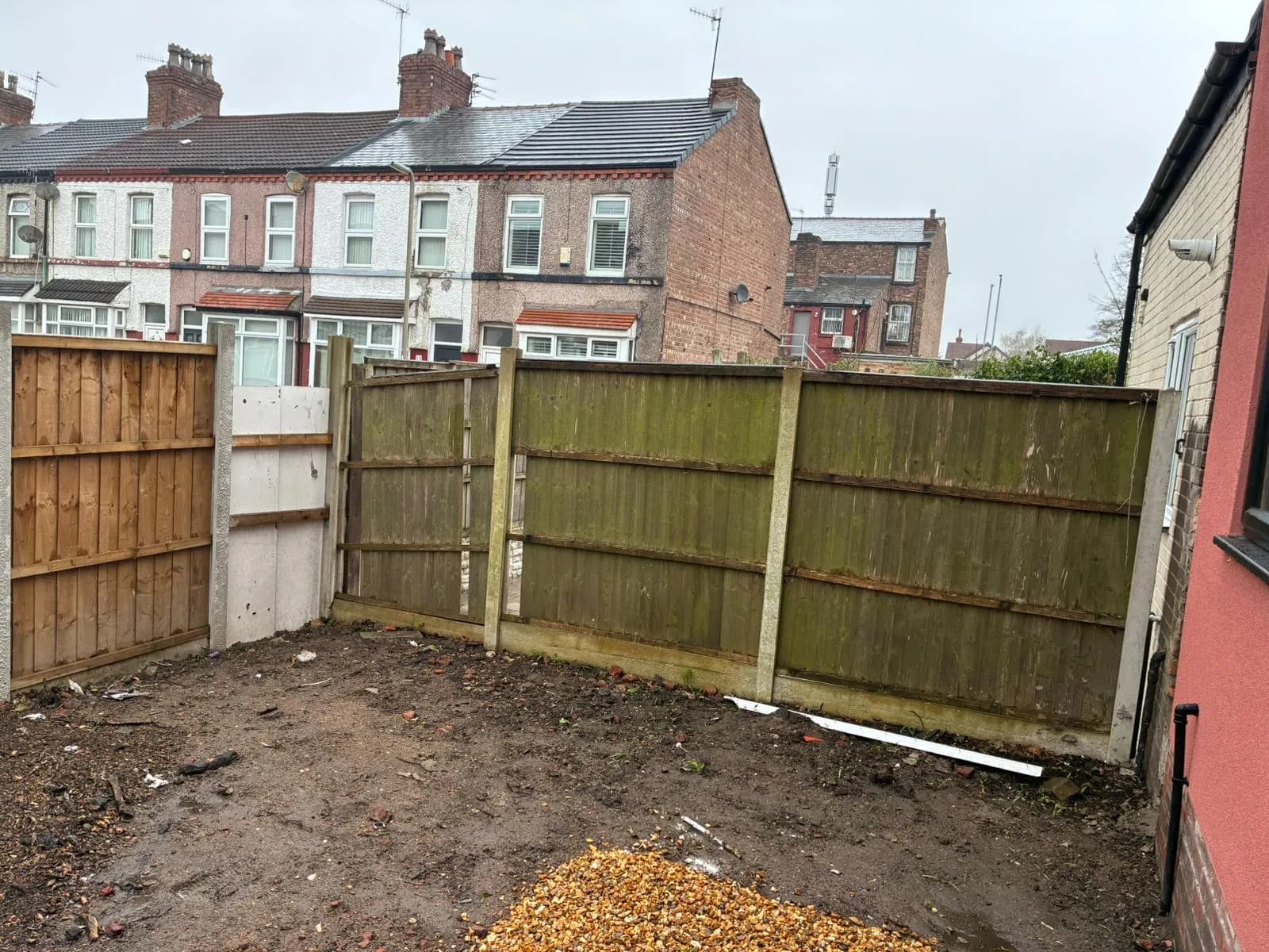 A rear yard dug out to soil, with an old lap panel fence on concrete posts and terraced houses behind
