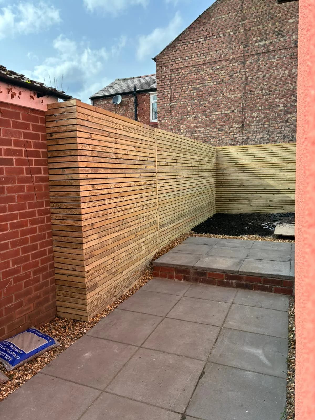 New horizontal slatted timber fencing on two sides of the yard, grey slab paving and a red brick step